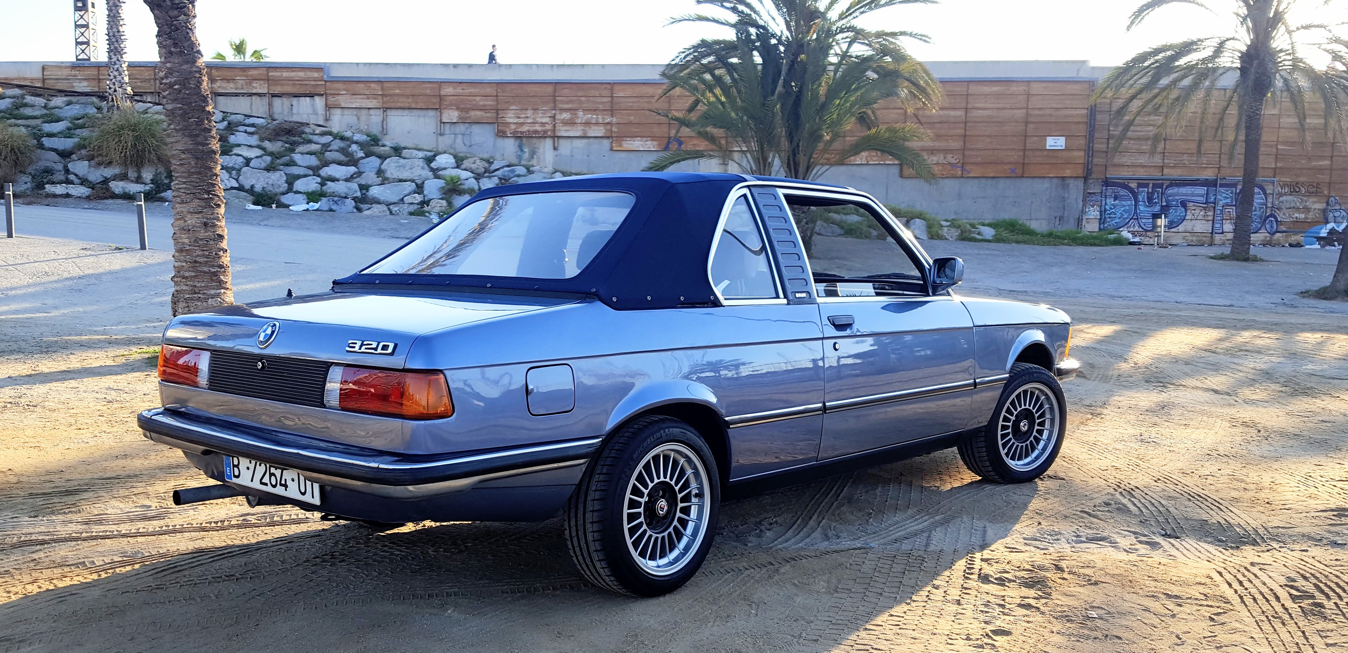 Rear three-quarter view of the BMW E21 on a Barcelona beach with palm trees — music video backdrop